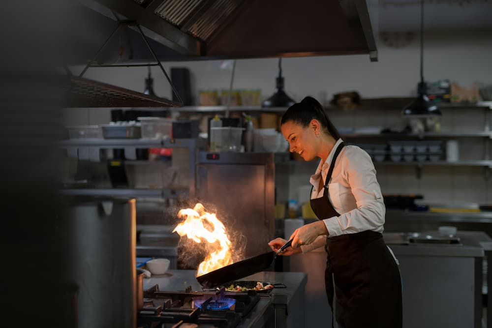 Una mujer en un restaurante profesional cocina con una sartén en llamas, demostrando habilidad en la gastronomía.
