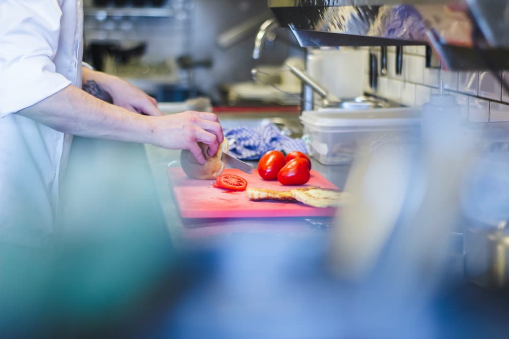 Una persona trabajando en una dark kitchen profesional, cortando un vegetal en una tabla de cortar roja. En la tabla también hay tomates y lo que parece ser pan. La cocina está equipada con utensilios, una estufa y recipientes de almacenamiento, representando el ambiente dinámico y práctico de este tipo de cocinas destinadas a la entrega de comida a domicilio.