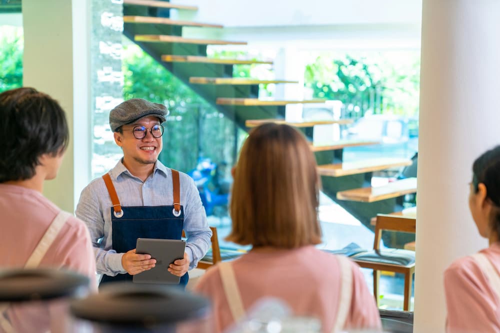 Chef sonriente en un café, usando un delantal y sosteniendo una tableta, conversando con el equipo en un ambiente moderno e iluminado.