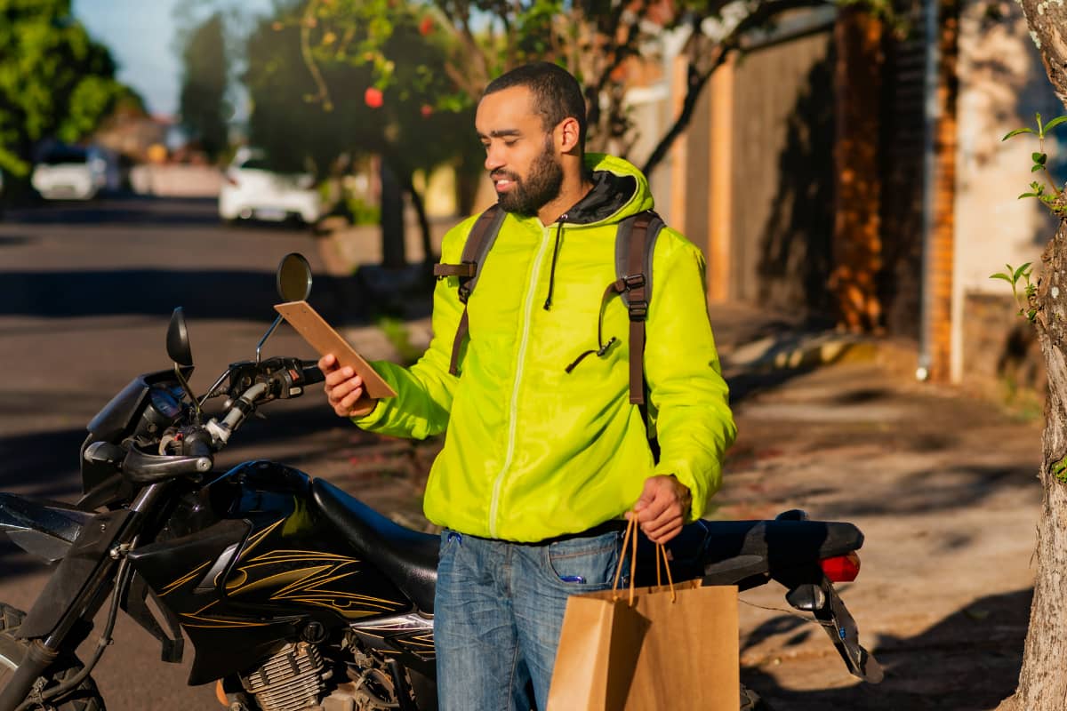 Entregador consultando pedido para reduzir o tempo de entrega do delivery, com sacolas e moto ao fundo em uma rua residencial.