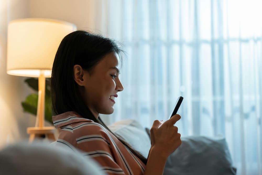 Mujer sonriente usando una app de delivery en un smartphone en la comodidad de la sala iluminada por una lámpara
