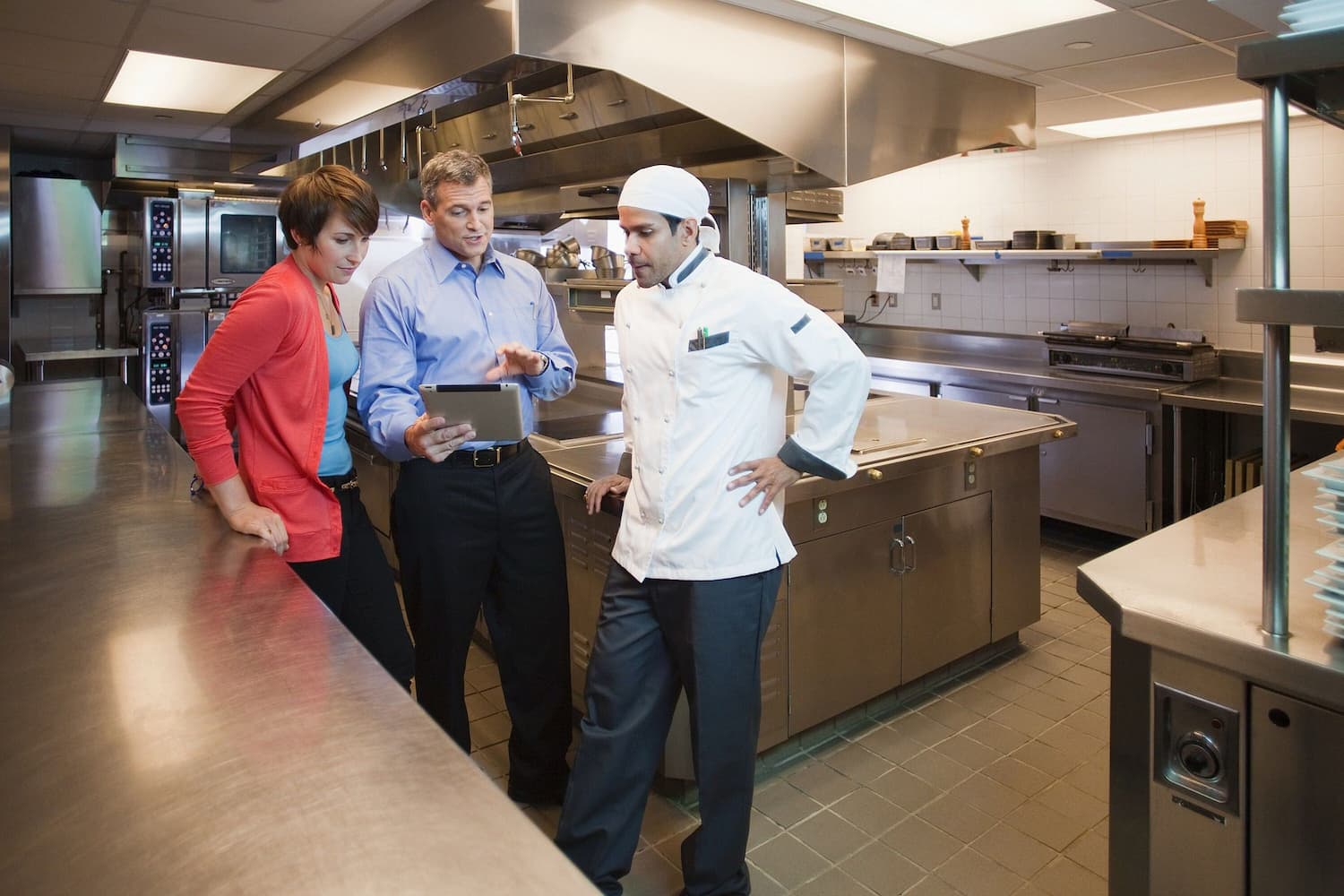 Três pessoas discutindo gestão de restaurante em uma cozinha industrial moderna. Uma pessoa está segurando um tablet enquanto conversa com um chef de cozinha e outra pessoa.