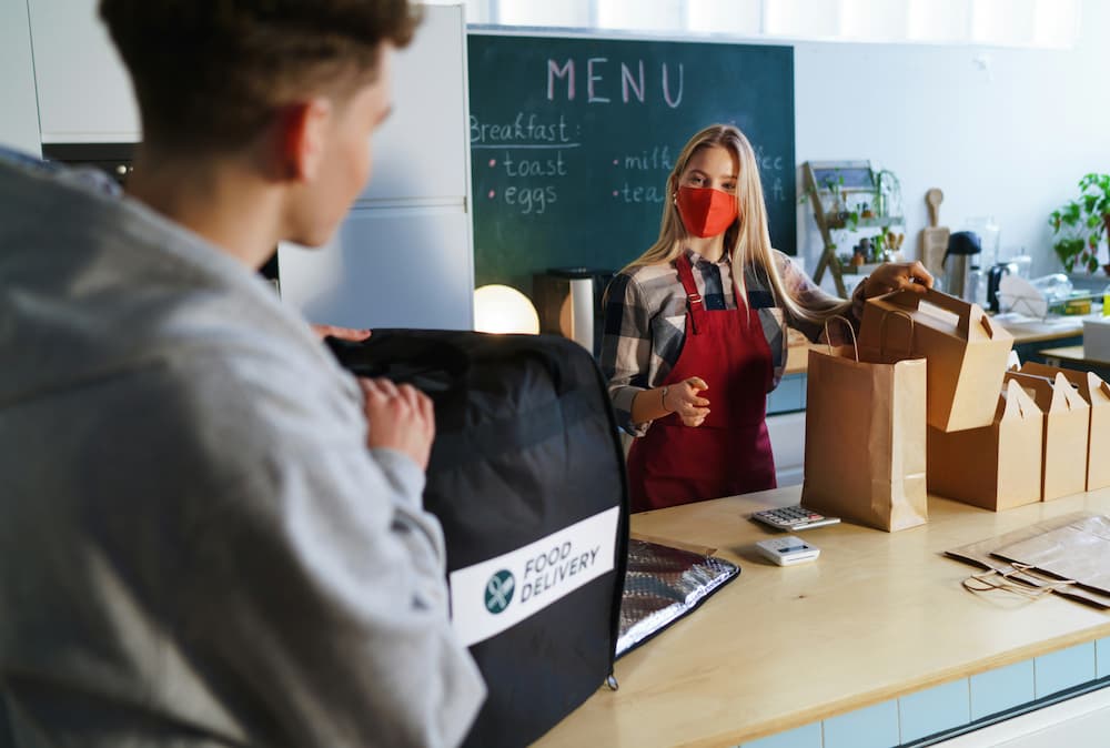 Pessoa realizando entrega de comida em um ambiente de cozinha, com sacolas e mochila de entrega visíveis