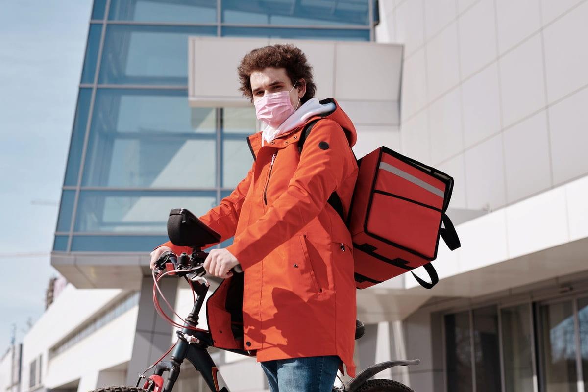 Repartidor de delivery en bicicleta con mochila térmica naranja, frente a un edificio moderno.