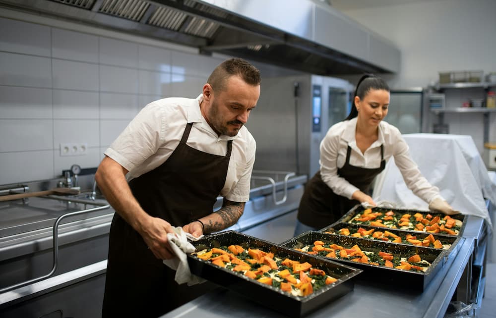 Dos chefs de cocina están organizando platos de verduras frescas y saludables en una cocina moderna.