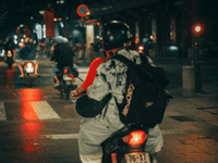 La imagen muestra a un repartidor nocturno conduciendo una motocicleta por una calle urbana iluminada por luces artificiales. Lleva casco y una mochila grande de estilo escolar que podría estar adaptada para entregas.