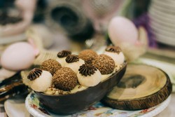 Ovo de Páscoa aberto, recheado com uma variedade de doces e trufas de chocolate, decorados com granulados coloridos. O ovo, feito de chocolate, está apresentado em um prato decorativo, com um fundo desfocado que inclui outros ovos decorativos, compondo uma cena atrativa e temática.