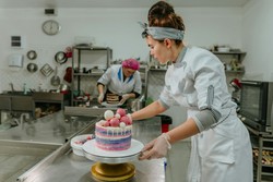 Confeiteira preparando um bolo colorido em uma confeitaria, com outra profissional ao fundo, criando sobremesas com criatividade e habilidade