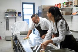 Dois funcionários estão higienizando uma bancada de aço inoxidável em uma cozinha industrial. Ambos vestem uniformes brancos e aventais pretos, reforçando a importância de boas práticas de higiene no ambiente de trabalho.