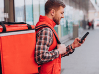 A imagem mostra um entregador de delivery parado em uma calçada, olhando para o celular.