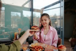 Uma pessoa vestindo uma camisa rosa está sentada em uma mesa de restaurante, cercada por diversos pratos, incluindo hambúrguer, batatas fritas, anéis de cebola e uma bebida. Outra pessoa está entregando um hambúrguer à pessoa sentada.