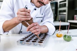 Confeiteiro profissional preparandando chocolates em forma de bombom na cozinha. O chef utiliza um saco de confeitar para moldar os doces com precisão usando chocolate derretido.