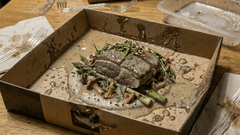 Um prato gourmet servido em uma caixa de apresentação escura sobre uma mesa de madeira clara. No centro, há um pedaço de carne coberto por um molho cremoso e ervas frescas, acompanhado de aspargos e cogumelos salteados.