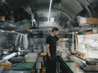 A imagem mostra um cozinheiro trabalhando em um ambiente compacto, moderno e altamente funcional — provavelmente dentro de uma cozinha móvel ou estrutura adaptada. Ele está preparando um prato com macarrão, cercado por ingredientes organizados em recipientes, equipamentos industriais e utensílios prontos para uso.