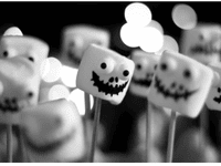 La imagen muestra varios malvaviscos decorados con caras espeluznantes de estilo Halloween. Cada malvavisco está montado en un palillo y presenta ojos saltones y bocas negras dibujadas con expresiones aterradoras o divertidas.