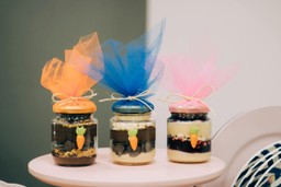 Três potes de vidro decorados como bolos de Páscoa, recheados com camadas de sobremesas, como chocolate, creme e frutas. As tampas dos potes são feitas de tecido colorido (laranja, azul e rosa) e estão amarradas com barbante, com pequenos enfeites de cenouras na frente.