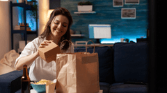 Sentada em um sofá azul-escuro, ela sorri enquanto retira uma caixinha de papelão de dentro de uma sacola de papel de delivery. À sua frente, há uma tigela com batatas chips e uma garrafa de cerveja, com a tela de um computador levemente desfocada ao fundo.
