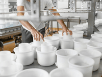 A imagem mostra uma pessoa trabalhando em uma linha de produção com vários recipientes brancos cilíndricos. O ambiente é limpo e organizado, sugerindo um processo industrial ou alimentício, com foco em produção em série.