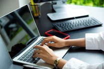 mão de mulher digita em notebook. Há uma mesa onde está um celular e teclado de computador