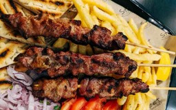 Três espetinho de carne acompanhados por batatas fritas, rodelas de tomate, fatias de pão e tiras de cebola.