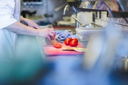 Una persona trabajando en una dark kitchen profesional, cortando un vegetal en una tabla de cortar roja. En la tabla también hay tomates y lo que parece ser pan. La cocina está equipada con utensilios, una estufa y recipientes de almacenamiento, representando el ambiente dinámico y práctico de este tipo de cocinas destinadas a la entrega de comida a domicilio.