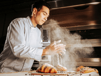 La imagen muestra a un panadero o pastelero en una cocina profesional, vestido con una chaqueta blanca, concentrado en su trabajo. Con las manos levantadas, está espolvoreando harina, que se dispersa en el aire y crea un efecto visual dinámico y expresivo.