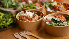 La imagen presenta una selección de comida saludable y fresca, servida en varios recipientes circulares de cartón ecológico sobre una mesa de madera clara. La composición sugiere un servicio de comida para llevar (takeaway)