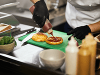 A imagem é um close-up focado na preparação de um hambúrguer em uma cozinha profissional, o que é altamente representativo do trabalho em uma dark kitchen (cozinha fantasma), onde a eficiência e a produção são centrais.