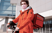 Entregador de comida parado com a sua bicicleta. Ele veste jaqueta laranja e tem uma caixa de transporte da mesma cor.