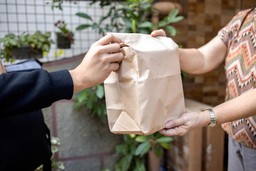 Braço de homem vestindo um moletom preto entregando uma sacola de papelão para uma mulher que veste uma blusa de manga curta estampada.