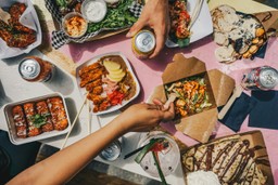 Una vistosa toma aérea (flat lay) de una mesa de madera pintada de rosa brillante, repleta de diversos platos de comida callejera internacional.