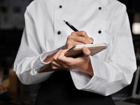 Chef de cozinha em uniforme branco com botões pretos e avental escuro escrevendo ou anotando em um bloco de notas espiral com uma caneta.