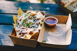 Deliciosos nachos com guacamole, cobertos com creme e pimenta, acompanhados de molho picante, servidos em uma embalagem de papelão para delivery