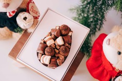 Ovo de Páscoa decorado com vários tipos de chocolates, colocado em uma bandeja branca sobre uma mesa de madeira. Ao redor, há dois ursinhos de pelúcia, um com roupa preta e laço vermelho segurando uma flor branca com etiqueta de "Love", e outro com roupa vermelha. Galhos verdes decorativos completam a cena ao fundo.