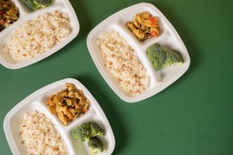 Prato de comida congelada com arroz, brócolis e frango temperado em recipientes de plástico em uma mesa verde.