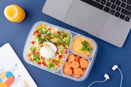 Imagem de marmita fitness com legumes, ovo cozido e molho, ao lado de um notebook e fone de ouvido, exemplificando como fazer marmita fitness para vender.