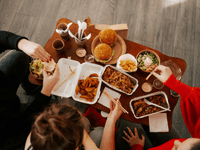 A imagem mostra um grupo de pessoas compartilhando uma refeição composta por diversos itens de comida por delivery dispostos sobre uma mesa de madeira. Entre os alimentos estão hambúrgueres, batatas fritas, saladas, pratos de macarrão asiático, legumes salteados, molhos em potes individuais e embalagens descartáveis.