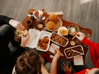 A imagem mostra um grupo de pessoas compartilhando uma refeição composta por diversos itens de comida por delivery dispostos sobre uma mesa de madeira. Entre os alimentos estão hambúrgueres, batatas fritas, saladas, pratos de macarrão asiático, legumes salteados, molhos em potes individuais e embalagens descartáveis.