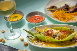 variedad de platos típicos de Semana Santa en Perú. En el centro, destaca un plato con choclo acompañado de una salsa amarilla, decorado con rodajas de ají y hierbas frescas. A la izquierda, se aprecia una copa de vino blanco junto a pequeños recipientes con salsas coloridas. Al fondo, hay otro plato que incluye pulpo y una guarnición de papas fritas finamente cortadas.