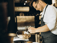 Descripción de la Imagen La imagen es una fotografía de un chef, de plano medio, en lo que parece ser una cocina profesional o un área de servicio, mientras está emplatando. 