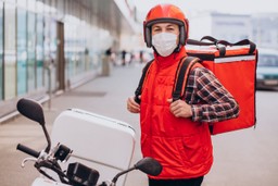 Delivery de comida: Motoboy vestido de colete laranja com uma blusa xadrez por baixo do colete com uma bag de entrega laranja, sua moto é branca, ele está na calçada da rua