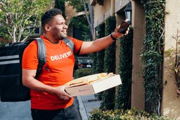 Homem segurando uma caixa e mochila de entrega preta, usando camiseta laranja &quot;delivery&quot; e calça jeans preta.