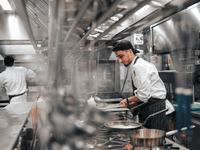 La imagen muestra a dos chefs trabajando en una cocina profesional con una atmósfera ajetreada y moderna.