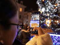 la imagen muestra a una persona sosteniendo un teléfono móvil durante la noche, tomando una fotografía de una escena iluminada por luces decorativas en un entorno urbano festivo. La toma está hecha desde atrás, dejando ver parte del rostro de perfil, con gafas, y la mano enfocada en la pantalla del celular.