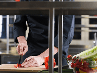 La imagen muestra a un chef en plena acción en una cocina profesional. El chef está vestido con un uniforme de cocina oscuro, posiblemente negro o azul marino, y sus manos están enfocadas mientras corta un pimiento rojo brillante sobre una tabla de cortar de madera clara