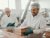 A imagem mostra um ambiente de cozinha industrial voltada para delivery, com dois profissionais usando roupas apropriadas de higiene: touca, jaleco branco e luvas. Um deles está sorrindo enquanto embala caixas de papelão, indicando um clima produtivo e organizado. Ao fundo, vê-se uma estrutura limpa, com equipamentos e prateleiras, típica de uma cozinha comercial moderna.