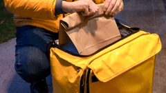 Esta imagen muestra a un repartidor de comida. El repartidor lleva una chaqueta amarilla de manga larga y pantalones vaqueros. Está agachado y sujetando una bolsa de papel marrón que contiene comida.