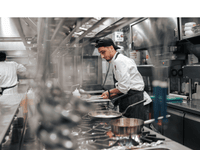 A imagem mostra o interior de uma cozinha industrial profissional. Em primeiro plano, um cozinheiro está trabalhando concentrado, usando uniforme branco com avental escuro e boné preto virado para trás.