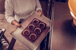 Uma pessoa vestindo um uniforme de chef está colocando uma bandeja de metal com uma forma de silicone contendo seis forminhas de papel com massa de bolo em um forno