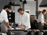 
A imagem mostra uma cena de trabalho concentrado em uma cozinha industrial ou profissional, com foco em três chefs ou cozinheiros.